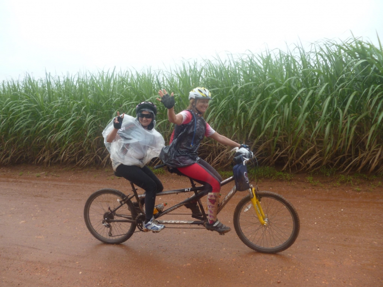 Imagem mostra Anabe conduzindo Ana Lídia por uma trilha, ambas sorridentes e acenando