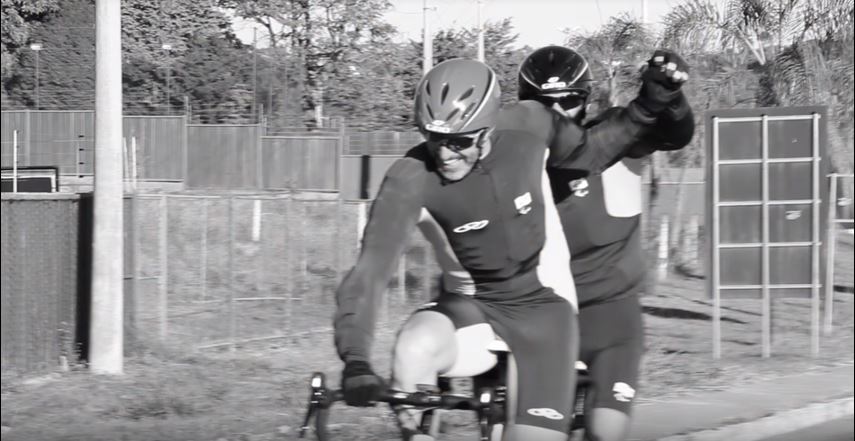 Imagem mostra, Weimar e Adauto, após completarem o desafio, ainda pedalando na tandem speed, com as mãos dadas em forma de vitória, comemorando o feito. 