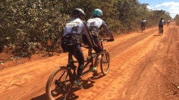 na imagem, uma dupla pedalando. Já passaram pela câmera e seguem ao encontro dos demais que estão à frente... Céu azul, chão de terra vermelha... coisas do cerrado